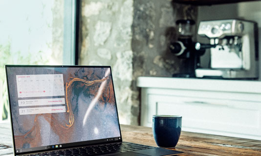 Photo of a laptop open on a table next to a cup with a coffee machine in the background